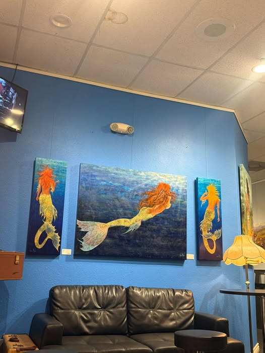 Three mermaid paintings by artist Jane Mick hanging on a blue wall inside The Main